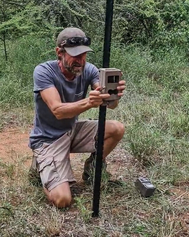 A man with a beard and cap kneels in a grassy field while attaching a trail camera to a vertical metal post.