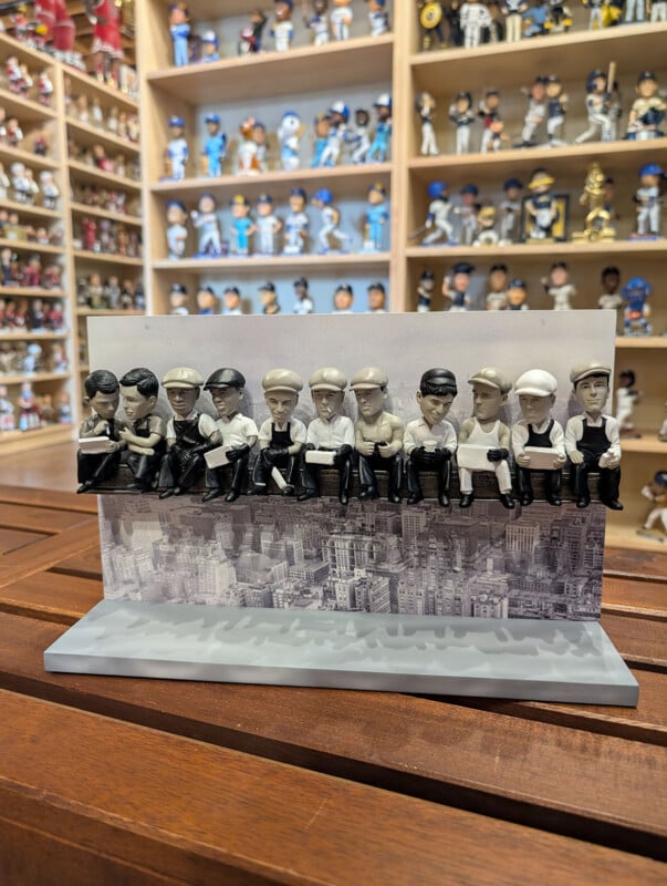 A figurine depicting the iconic "Lunch atop a Skyscraper" photograph sits on a wooden table in front of shelves filled with bobbleheads.