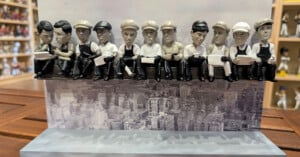 A bobblehead display featuring eleven figures sitting on a steel beam, modeled after the famous "Lunch Atop a Skyscraper" photograph, set against a backdrop of a city skyline.