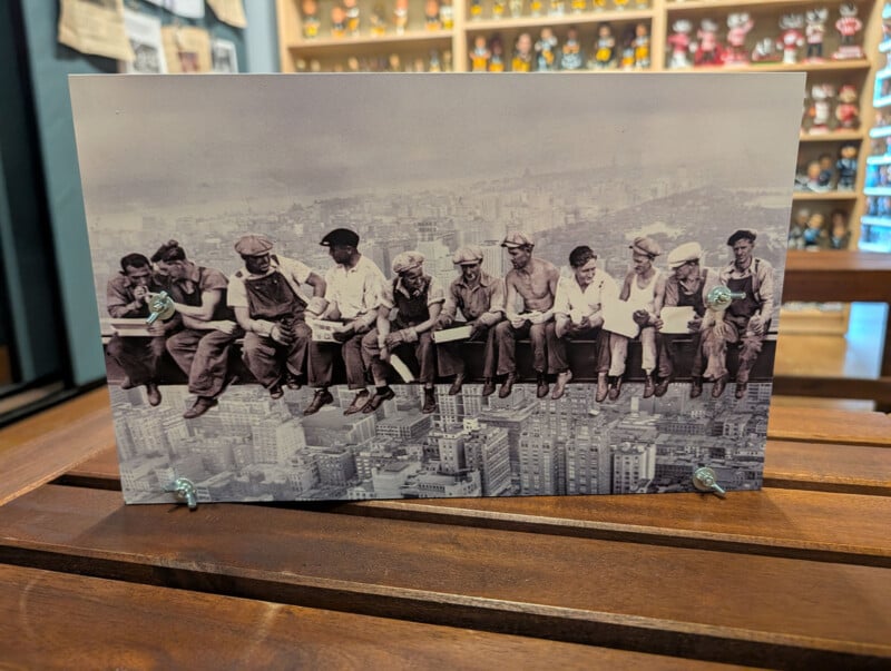 A black-and-white print of the iconic "Lunch atop a Skyscraper" photograph mounted on a wooden stand with four metal wing nuts.