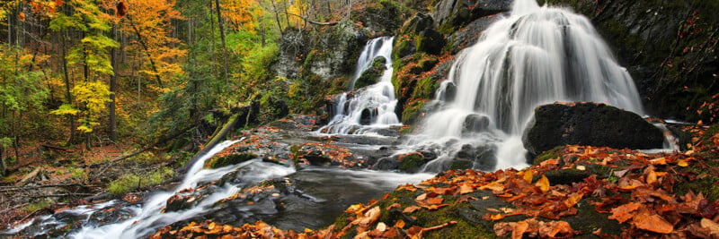 A waterfall cascades over dark, mossy rocks into a stream surrounded by a forest with vibrant autumn foliage.