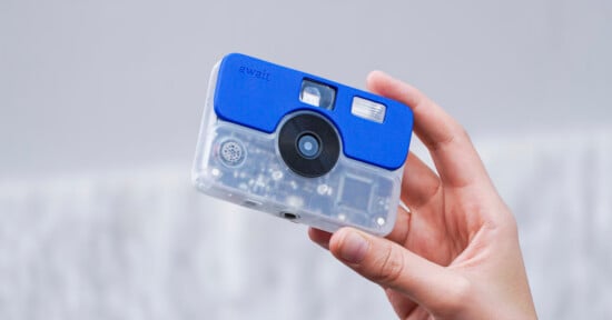 A hand holds a small, rectangular digital camera with a transparent body and a blue plastic top section labeled "await.