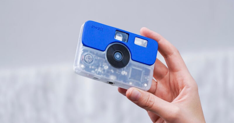 A hand holds a small, rectangular digital camera with a transparent body and a blue plastic top section labeled "await.