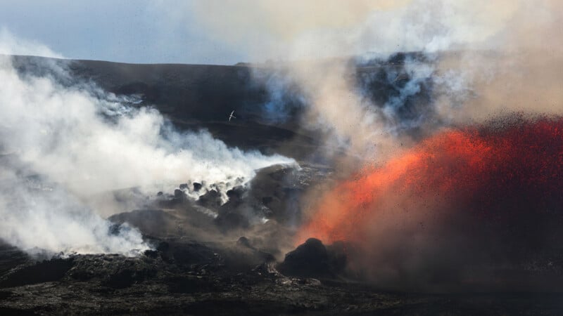 A volcanic eruption spews glowing red lava and dark ash into the air, with thick white smoke billowing across a rocky landscape.