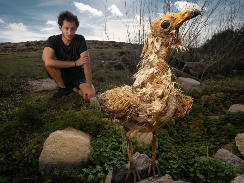 A man crouches on a grassy hillside next to a large, weathered, bird-like sculpture with textured, frayed plumage.