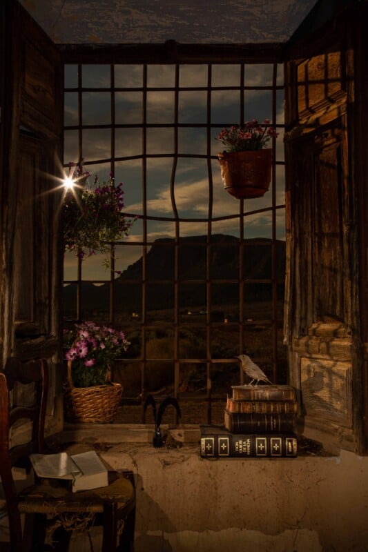 An open wooden window with iron bars overlooks a mountain landscape at sunset, featuring a chair with an open book, potted flowers, and a stack of books topped with a bird figurine.