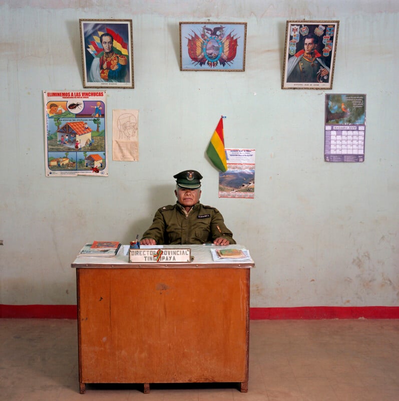 A man in a military uniform sits behind a wooden desk labeled "Director Provincial Tingipaya" in a room decorated with portraits, a flag, and posters.