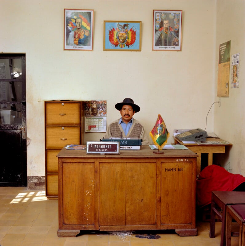 A man in a hat sits at a wooden desk labeled "INTENDENTE BETANZOS" in an office with portraits of Simón Bolívar and Antonio José de Sucre on the wall.