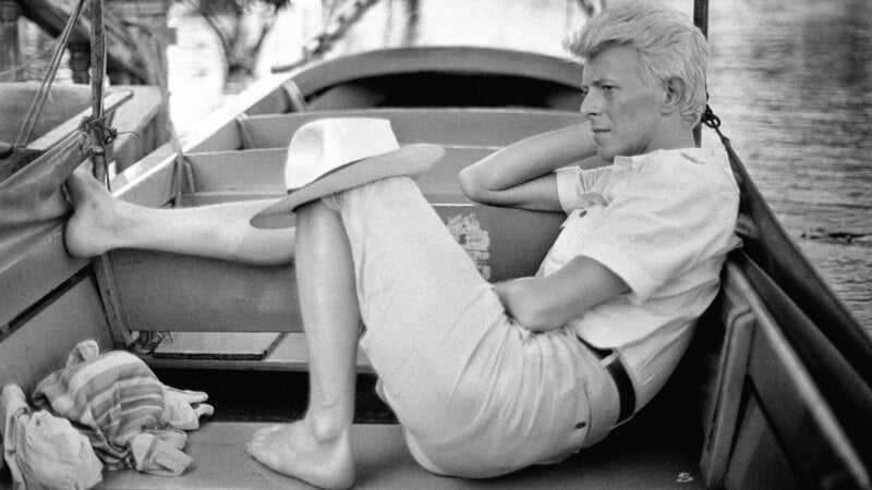 David Bowie sits barefoot in a wooden boat, wearing a light-colored shirt, trousers, and a hat resting on his knee.