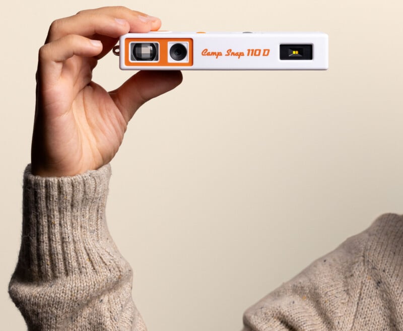 A hand in a beige sweater holds a white and orange Camp Snap 110 D digital camera against a plain light background.