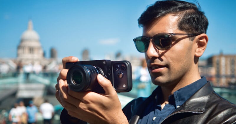 A man wearing sunglasses and a leather jacket holds a smartphone attached to a large camera lens, with a domed building blurred in the background.