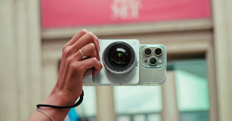 A hand holds a smartphone attached to a large, circular camera lens attachment, with a wrist strap visible on the arm.