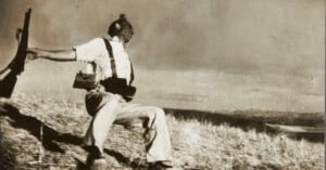 A sepia-toned photograph shows a soldier in a white shirt and trousers falling backward on a grassy hillside, his rifle slipping from his outstretched hand.