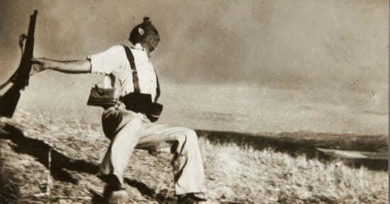 A sepia-toned photograph shows a soldier in a white shirt and trousers falling backward on a grassy hillside, his rifle slipping from his outstretched hand.