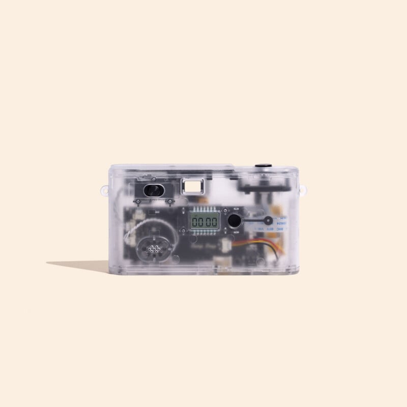 A clear, transparent digital camera with visible internal circuitry, a small LCD screen displaying "00 00," and a flash unit, set against a light beige background.