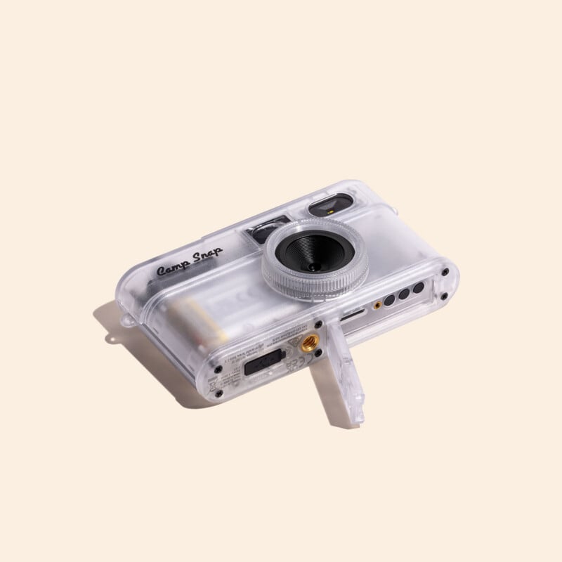 A clear, translucent digital camera with the text "Camp Snap" on the front, shown at an angle with its bottom port cover open.