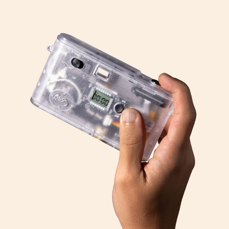 A hand holds a clear, transparent film camera against a light beige background, showing the internal components and a small digital display.