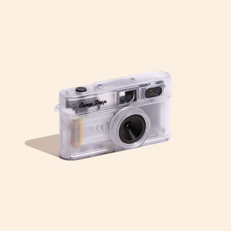 A clear, translucent digital camera with a black shutter button and a circular lens sits on a light beige background.