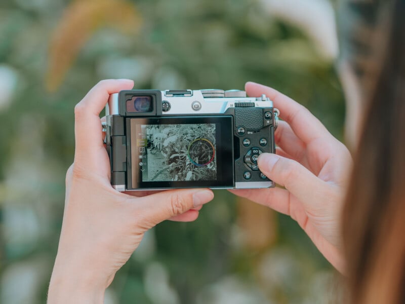 A person holds a digital camera, displaying a monochrome image with a color adjustment wheel on its screen, against a blurred green background.