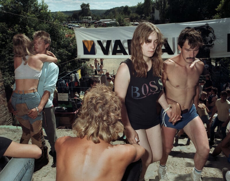 A group of young people in casual summer clothing gather outdoors near a banner, with some individuals embracing or walking together.