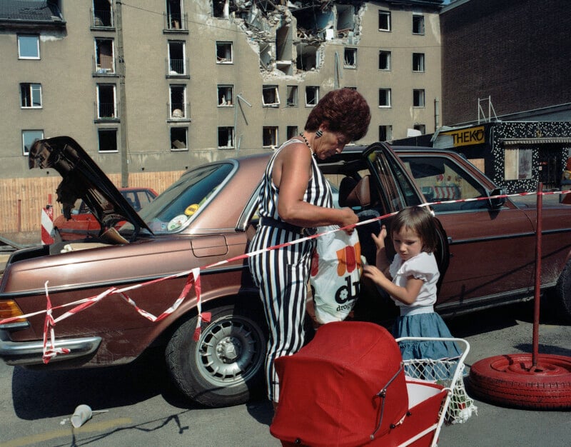 A woman in a striped jumpsuit and a young girl stand by a brown Mercedes-Benz parked in front of a damaged, partially collapsed building.