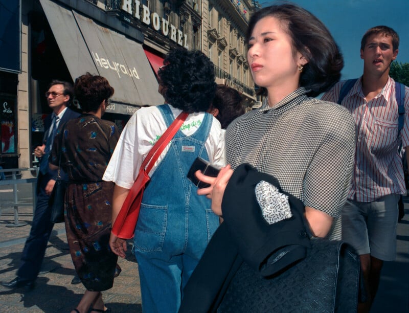A woman in a checkered blouse walks through a crowded city street, with pedestrians and storefronts visible in the background.