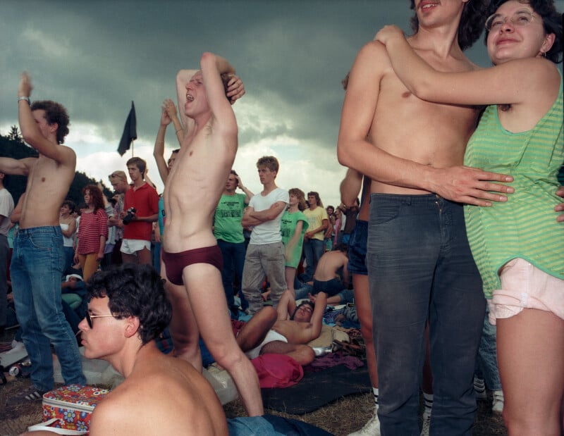 A crowd of young people at an outdoor festival, with some standing and others sitting on the grass under a cloudy sky.