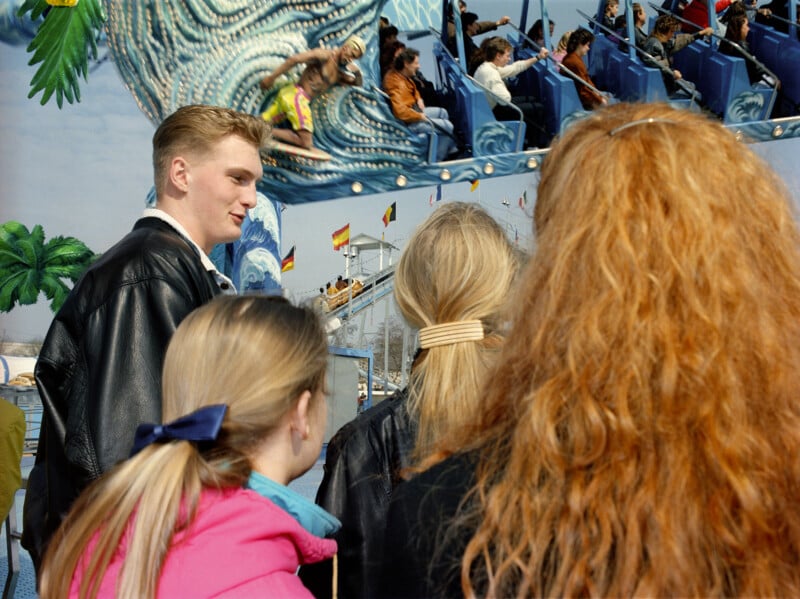 A young man in a leather jacket stands with three women, all facing a fairground ride featuring a painted surfer.