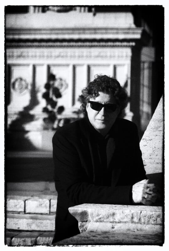 A man with curly hair and sunglasses wearing a dark jacket leans against a stone ledge in front of a classical building.