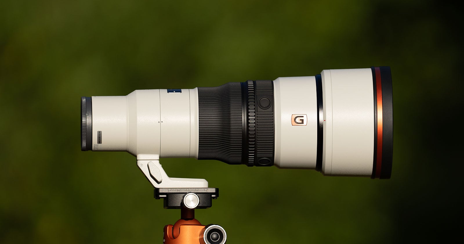 A white telephoto camera lens with a black focus ring and a G logo is mounted on a tripod head against a blurred green background.