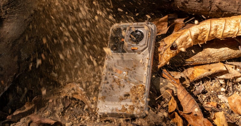 A rugged smartphone sits on the ground among dry leaves and dirt, partially obscured by a spray of flying debris.