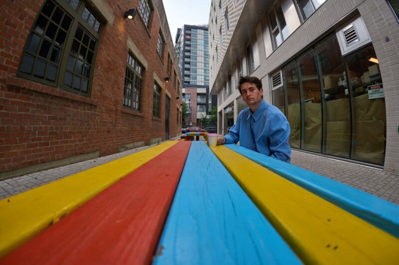A man in a blue button-down shirt sits at a brightly painted yellow, red, and blue picnic table in an urban alleyway.