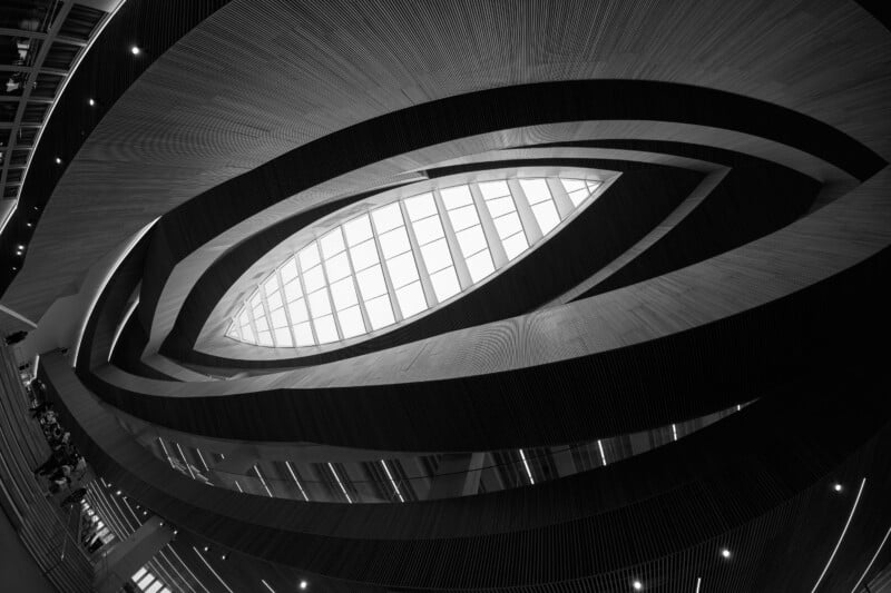 A low-angle, black-and-white view of a modern atrium with curved, slatted wooden balconies surrounding a central, eye-shaped skylight.