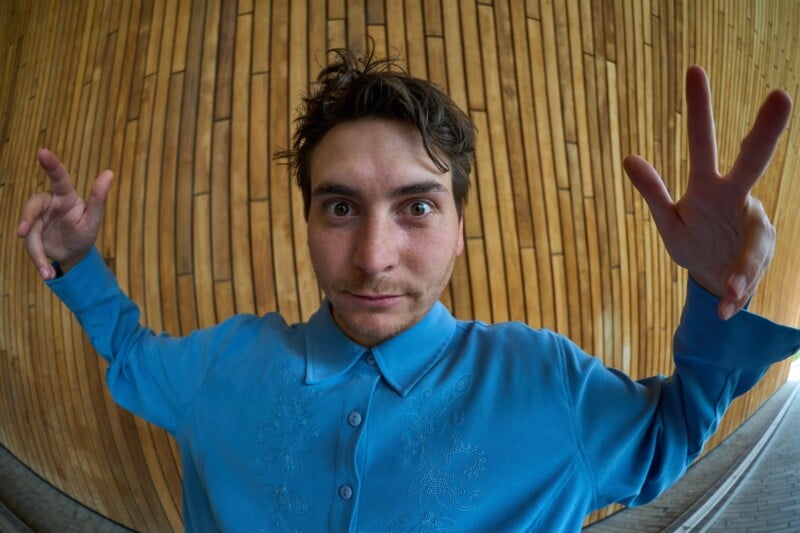 A person with messy brown hair wearing a blue embroidered button-down shirt poses with both hands raised in front of a curved wooden wall.