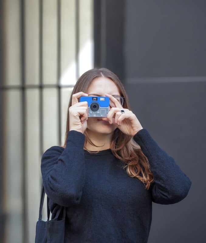 A woman with long brown hair wearing a black sweater holds a small blue and clear film camera up to her eyes to take a photo.
