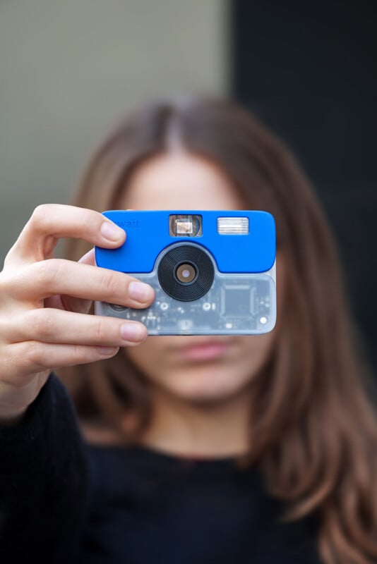 A person holds a small, blue and translucent digital camera in front of their face, obscuring their features.