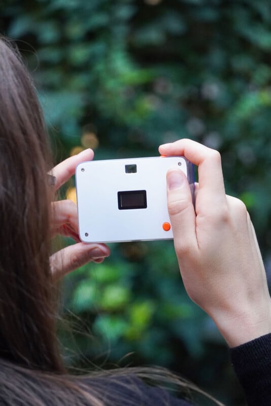 A person with long brown hair holds a small, white rectangular digital camera with an orange button, aiming it toward a blurred green background.
