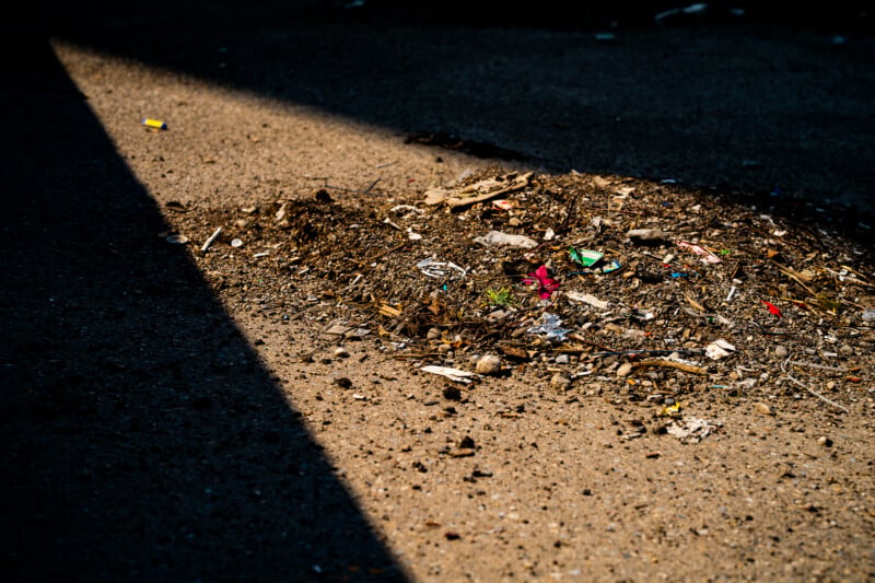 A patch of dirt and scattered debris is illuminated by a sharp beam of sunlight against a dark, shadowed background.