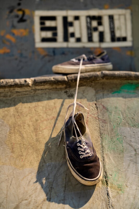A pair of dark blue canvas sneakers are tied together by their laces, with one resting on a ledge and the other hanging down against a textured, graffiti-covered wall.