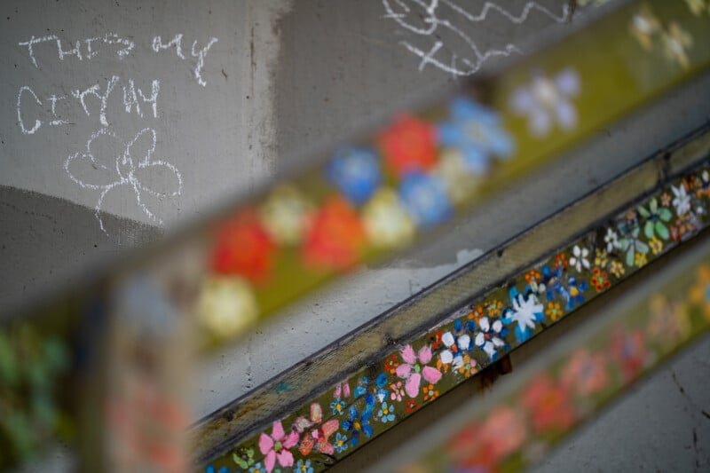 A concrete wall with chalk graffiti reading "This is my city" next to a hand-drawn flower, partially obscured by a blurred, flower-painted wooden bench in the foreground.