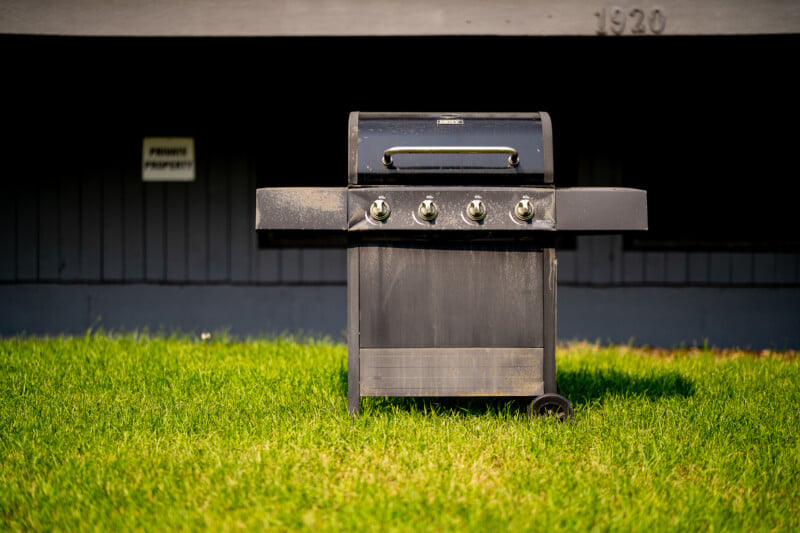 A black gas grill stands on a green lawn in front of a gray building with the number 1920 and a small sign visible on the wall.