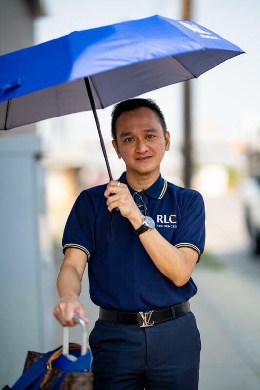 A man in a navy blue polo shirt with an RLC Residences logo holds a blue umbrella while standing next to a piece of luggage.