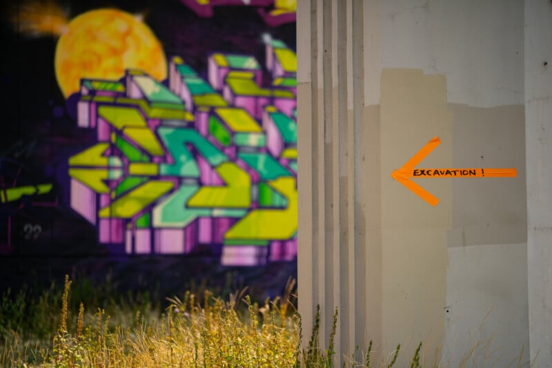 An orange tape arrow pointing left with the word "EXCAVATION!" written on it is attached to a concrete pillar next to a graffiti-covered wall.