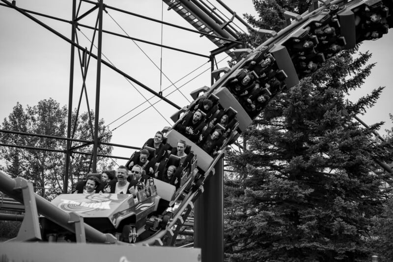 A black-and-white shot of a roller coaster train filled with passengers ascending a steep track next to a large evergreen tree.