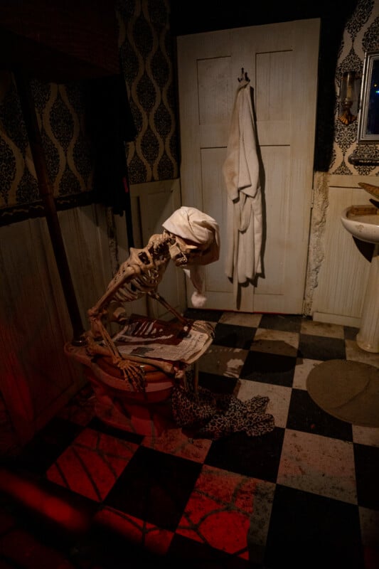 A skeleton wearing a nightcap sits on a toilet in a bathroom with checkered floors, reading a newspaper while a white robe hangs on the door.