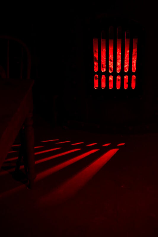 A dark room is illuminated by a red light source behind a vertical-slatted grate, casting long, parallel red shadows across the floor near a wooden table leg.