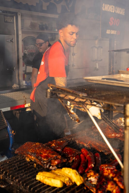A man in a red shirt and black apron grills ribs, sausages, and pineapple slices on a smoky outdoor barbecue grill.