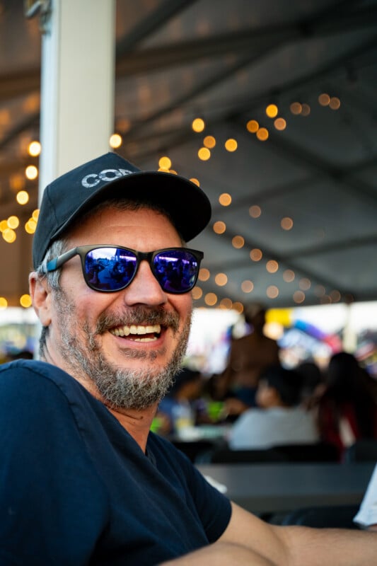 A smiling man with a beard and a black baseball cap wears reflective blue sunglasses while sitting under a tent with string lights.
