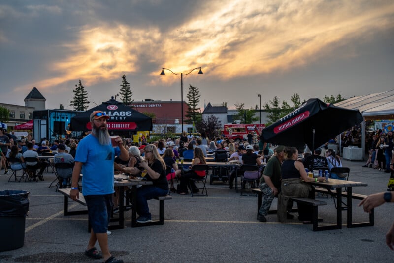 A man with a long beard walks through an outdoor festival area filled with people sitting at picnic tables under umbrellas at sunset.