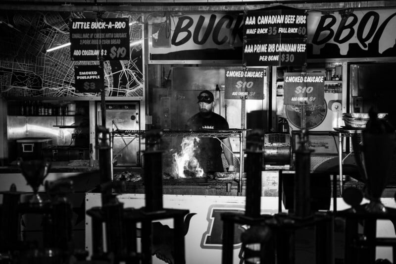 A man in a baseball cap grills meat over an open flame at a barbecue stand with menu signs hanging above.